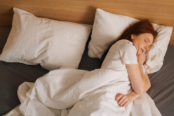 Top view of cute calm redhead young woman wearing soft comfortable night clothes sleeping in comfortable bed lying down on soft pillow. Peaceful pretty lady resting covered with blanket.