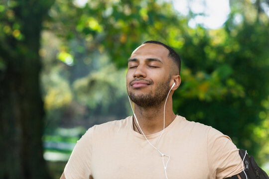 Close-up portrait of sportsman in park, hispanic man jogging in park with eyes closed breathing fresh air and resting, jogging with headphones listening to music and online radio and podcasts.