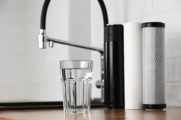 A glass of clean fresh water and set of filter cartridges on wooden table in a kitchen interior. Installation of reverse osmosis water purification system. Concept Household filtration system.