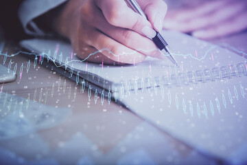 A woman hands writing information about stock market in notepad. Forex chart holograms in front....