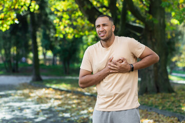 Naklejka na ściany i meble Hispanic athlete in the park has severe chest pain, man has a heart attack while jogging and doing fitness and exercise in the park outdoors.