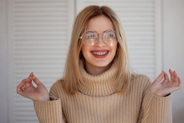 Stylish and beautiful young student with glasses and beige oversize sweater. Young woman wearing braces and smiling - obrazy, fototapety, plakaty