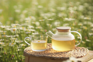 Beautiful nature landscape with chamomile field and small wooden table with camomile hot tea in transparent teapot and glass cup. Copy space