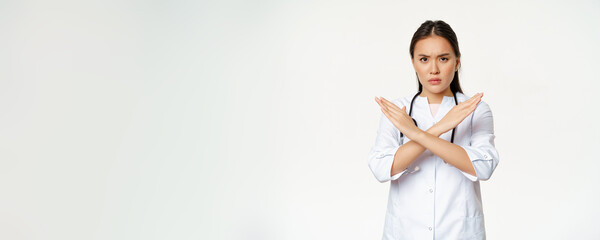 Serious asian woman doctor prohibit patient behaviour, showing stop, cross arms gesture, frownign displeased, disapprove smth bad, standing over white background