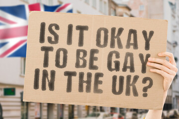 The question " Is it okay to be gay in the UK? " is on a banner in men's hands with blurred background. Friendly. Passionate. Contact. Date. Dating. Lover. Partner. Boyfriend. Pleasant. Approval