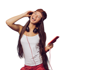 Charming young brunette girl with long hair dressed in white t-shirt, baseball cap and red pants, listening music with phone, headphones on her head, shows rock hand gesture over transparent backdrop.