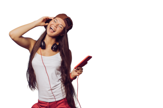 Charming young brunette girl with long hair dressed in white t-shirt, baseball cap and red pants, listening music with phone, headphones on her head, shows rock hand gesture over transparent backdrop.