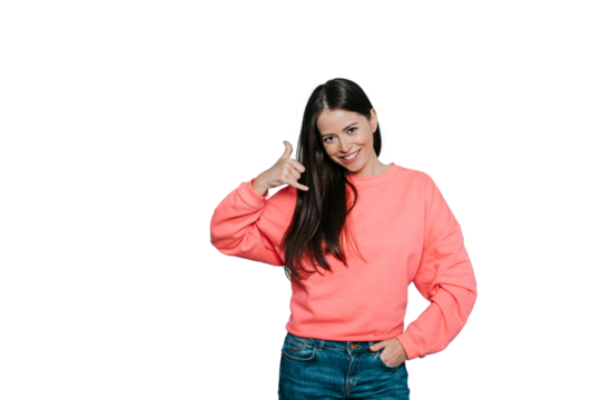 Smiling delighted brunette woman makes phone gesture, shows call me back sign with hand near ear, pretends talking on mobile phone, dressed in pink jumper, over transparent background.