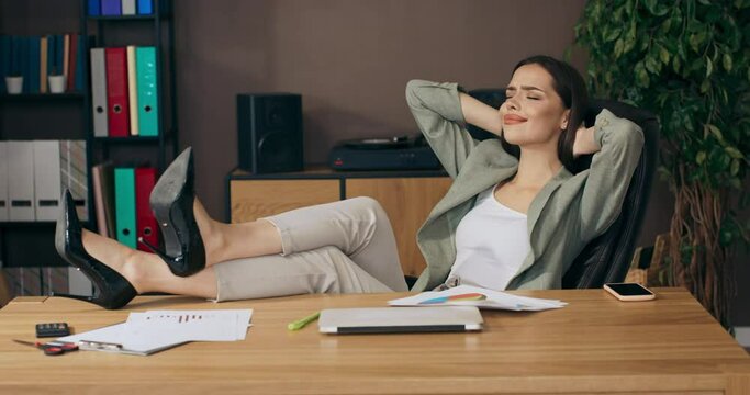 Calm beautiful adorable woman sitting at table in office holds legs on table relaxes after hard working day at home online job dream life for lady.