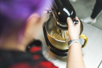 Pastry chef POV. Woman with colorful hair using mixer standing at counter in her apron. Professional bakery kitchen. High quality photo