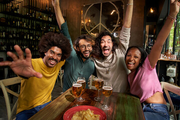 Group of four cheerful young friends taking selfie portrait inside the bar at happy hour. Happy people having fun looking at the camera smiling. Concept of community, youth lifestyle and friendship.