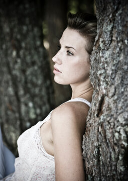 A young woman in a white tank top rests with her back against a tree.