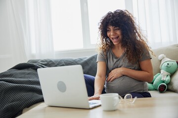 Pregnant woman smile sits at home on the sofa with a laptop and talks on video chat, blogger, freelancer works online. Home clothes, lifestyle of a pregnant woman, preparation for childbirth