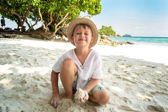 Little happy boy in summer hat playing with sand on the tropical beach, looking at the camnera with big smile on his face. Beautiful landscape. Blue sky and sea water background.