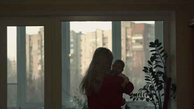 An elderly mother soothes her crying baby, in her arms, standing near window during air raid alert. Against background high-rise building, high-rise building, during day, winter, autumn