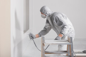 Decorator in protective overalls painting wall with spray gun indoors - obrazy, fototapety, plakaty