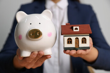 Woman holding piggy bank and little house model on light grey background, closeup