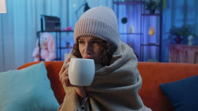 Sick woman wrapped in plaid sit alone shivering from cold on sofa drinking hot tea in unheated apartment without heating due debt. Unhealthy ill girl feeling discomfort try to warm up at evening home