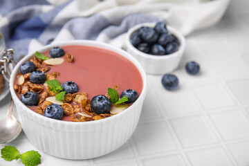 Bowl of delicious smoothie with fresh blueberries and granola on white tiled table, closeup. Space for text