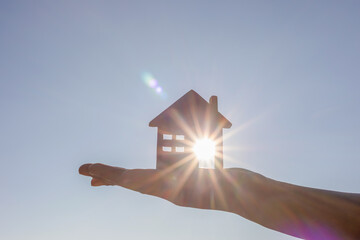 Hand holding wooden house toy against blue sunny sky with sun rays. solar power.