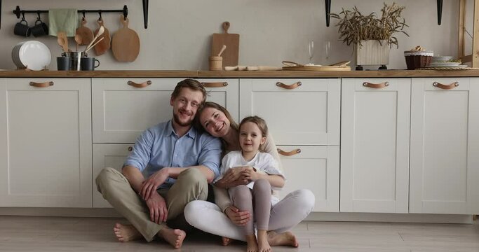 Young loving couple and lovely little 5s preschooler daughter posing looking at camera seated on warm floor in modern renovated kitchen. Happy young homeowners family portrait, bank loan and tenancy
