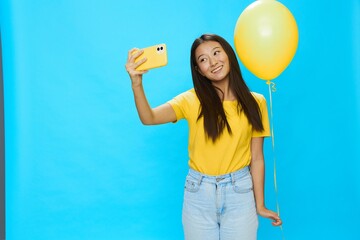 Fototapeta premium Asian woman holding the phone and looking at the screen talking on a video call with a yellow balloon in her hands on a blue background in a yellow T-shirt smiling with teeth 