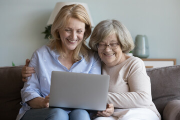 Joyful happy adult daughter and elderly mom resting on home sofa together, using laptop computer for communication, shopping with online service, Internet app, enjoying family leisure