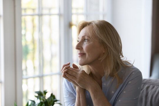 Positive thoughtful pretty middle aged blond woman sitting on home sofa, keeping hands at chin, looking at window away with good thoughts, thinking over future retirement, planning, dreaming