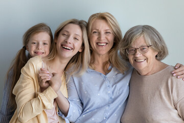 Happy little child, cheerful young mom, middle aged granny, elderly great grandma portrait. Four girls and women of different generations standing at grey wall background, hugging with love, smiling