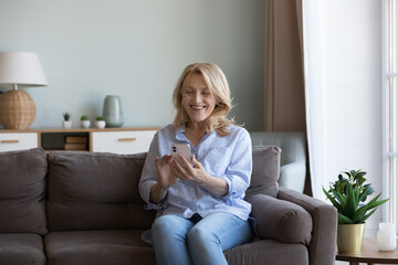 Happy retired mature woman smiling at cellphone display, using mobile phone at home for Internet communication, online chat, typing, touching screen, laughing, sitting on sofa, holding gadget