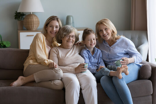 Happy joyful daughter kid, mom. grandma, elderly great grandmother meeting at home, resting on sofa, hugging, holding cups, enjoying leisure, family party, looking at camera, smiling