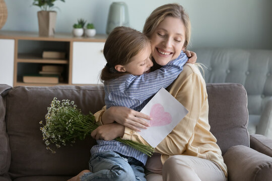 Happy peaceful young mom hugging adorable daughter with love, gratitude, holding flowers, greeting card with drawn heart, smiling with closed eyes, laughing, celebrating mothers day