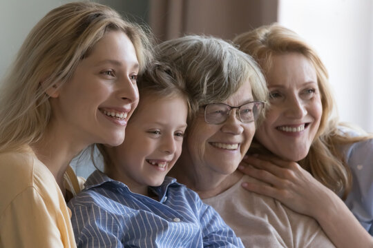 Happy pretty girls and mature senior women of four generations sitting close together, posing, looking away with toothy smiles, laughing, hugging, enjoying family leisure time