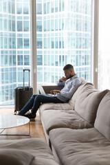 Millennial serious pensive African businessman sit on sofa working on laptop stay in luxury hotel on business trip, skyscraper buildings city view through panoramic window, vertical shot. Lifestyle