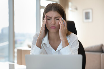 Frustrated woman working at office desk in front of laptop touching temples, suffers from chronic headache due to information overload, electromagnetic radiation, closed eyes, relieving pain, migraine