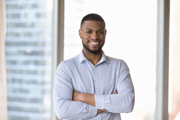 Fototapeta Millennial successful businessman standing in modern skyscraper office smile staring at camera looks confident. Portrait of business owner, company boss pose with arms crossed on chest at workplace obraz