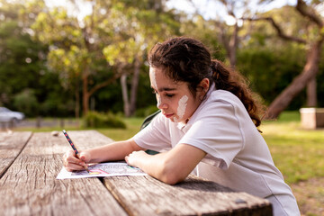 Young Aboriginal girl with white face paint sitting at an outdoor table drawing