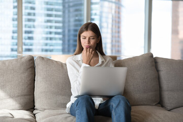 Frowned young woman sit on sofa at home staring at laptop screen having dilemma, doubts or...