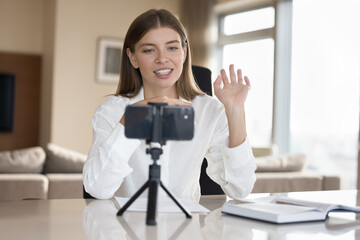 Attractive woman greets followers sit at desk staring at cellphone screen start streaming, making video call by study, filming job interview. Modern tech usage for distancing communication, vlogging