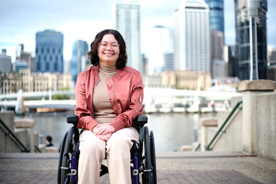 Smiling woman with a disability sitting in a wheel chair with tall buildings behind her