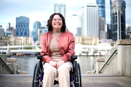 Smiling woman with a disability sitting in a wheel chair with tall buildings behind her