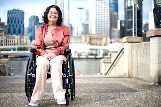 Smiling woman with a disability sitting in a wheel chair with tall buildings behind her