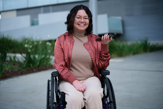 Smiling woman with a disability wearing glasses sitting in a wheel chair holding a mobile phone