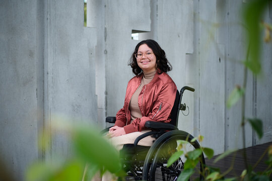 Smiling woman with a disability sitting in a wheelchair outside in urban setting with greenery