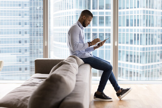 Busy African businessman work on digital tablet sits on sofa in modern skyscraper office lobby or living room at home. E-business, marketplace or business application user, electronic services buyer