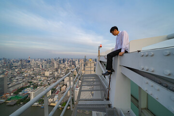 Depressed lonely Asian businessman is fired from job. Sitting on rooftop with Bangkok urban downtown city background, Thailand. People lifestlye. Danger. Unhappy life. Suicide