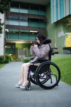 Woman in facemask with disability in a wheelchair outside looking at her mobile phone