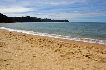 View of a golden sand beach with blue sky and ocean water, and native bush-covered mountains in the background.