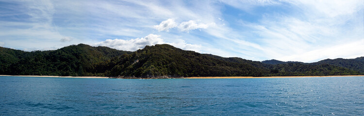 Fototapeta premium View of a golden sand beach with blue sky and ocean water, and native bush-covered mountains in the background.