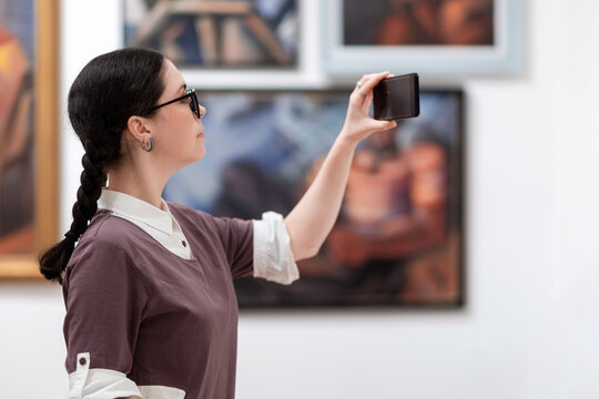 Cultural education and modern exhibition in gallery. Side view of woman visiting excursion in museum. Student using smartphone and take photo. Concept of Museum's day
